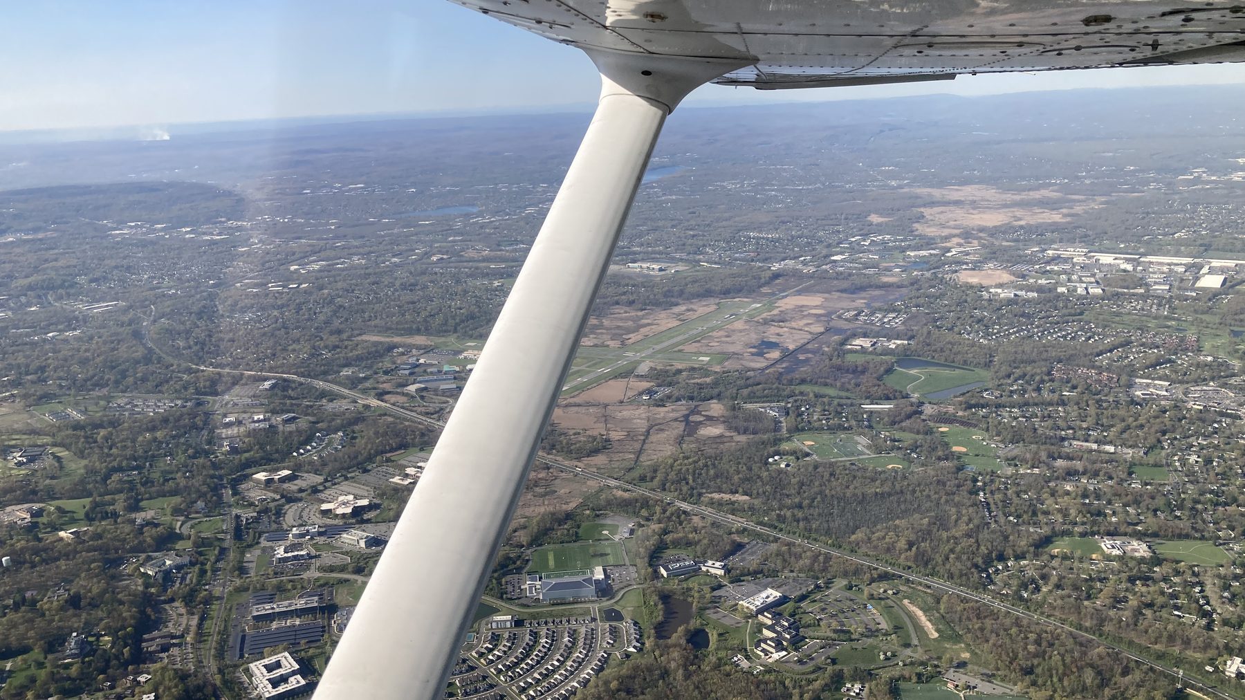 Wing and strut of a Cessna over suburban New Jersey, a general aviation airport visible below.