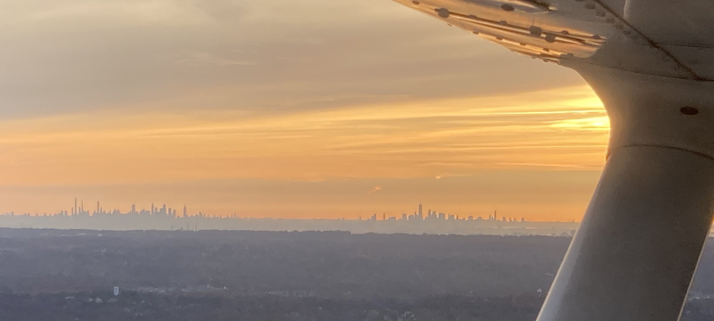 Golden-hour view past the wing strut of a training aircraft, with the Manhattan skyline on the horizon.