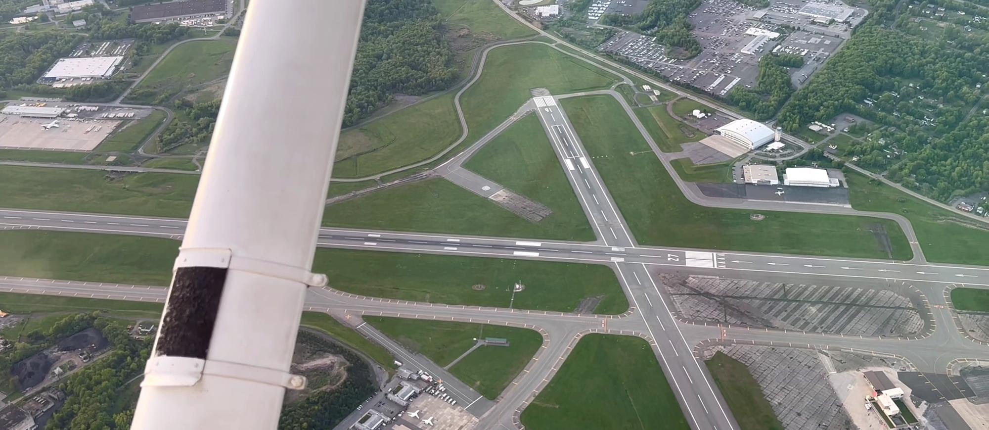 An airport's crossing runways and taxiways seen past the wing strut of a training airplane on downwind.