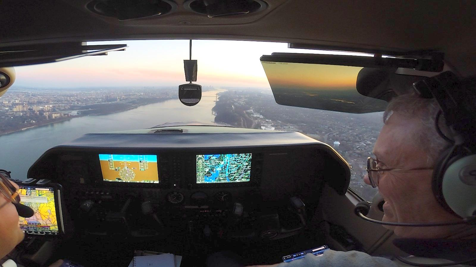 Flight instructor and student in the cockpit of a training aircraft over the Hudson River at dusk, with the George Washington Bridge and the Manhattan skyline ahead.